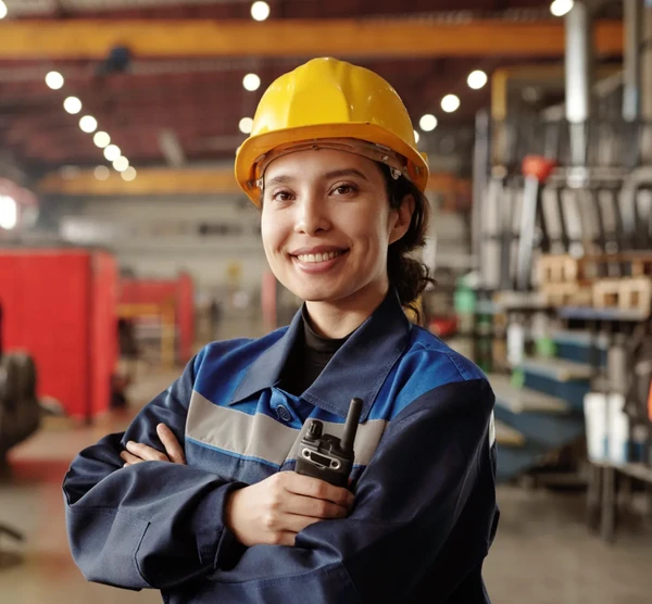 Smiling worker in a yellow hard hat and blue uniform holds a walkie-talkie in an industrial setting.