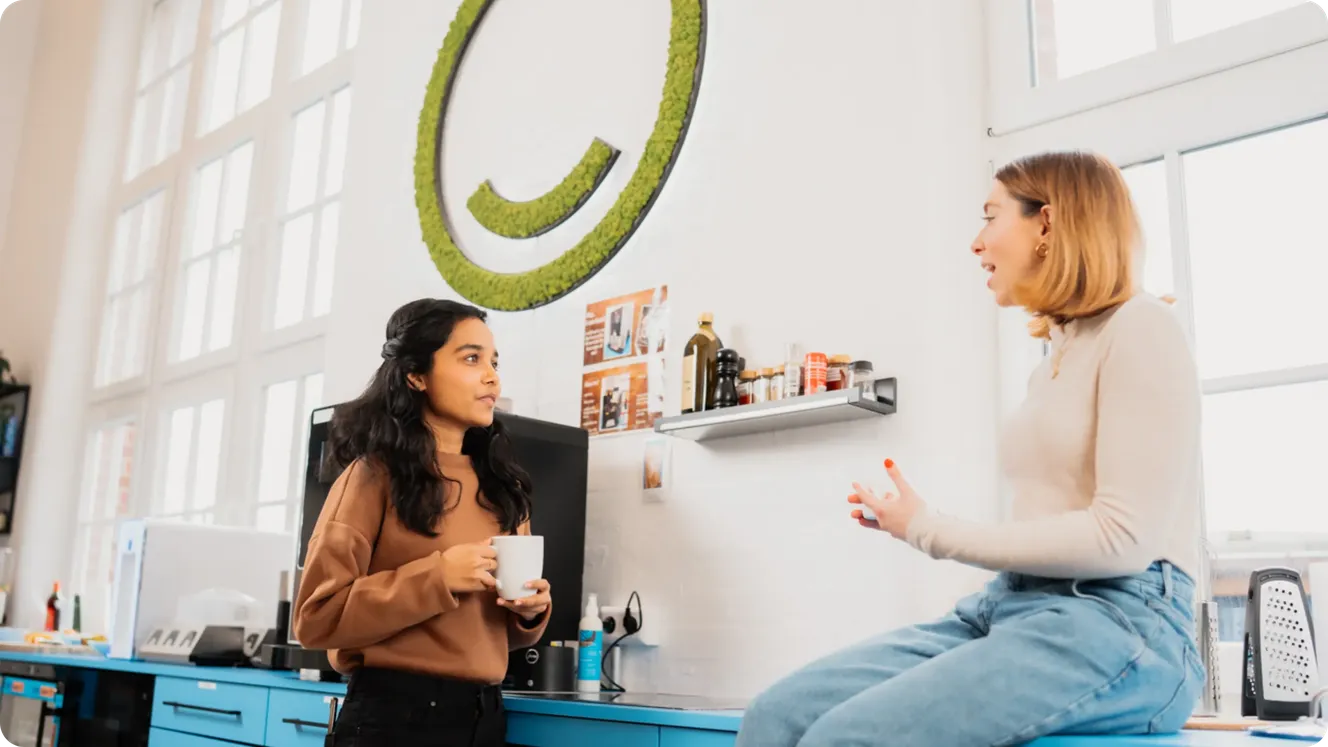 Two Staffbase colleagues chatting over coffee in the office kitchen beneath a green moss logo.
