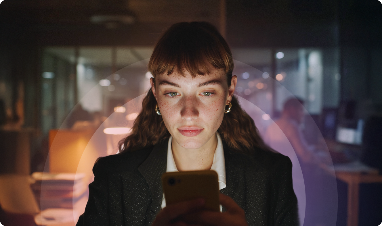 Woman in a dark office focused on her smartphone, with a soft light illuminating her face and blurred background.