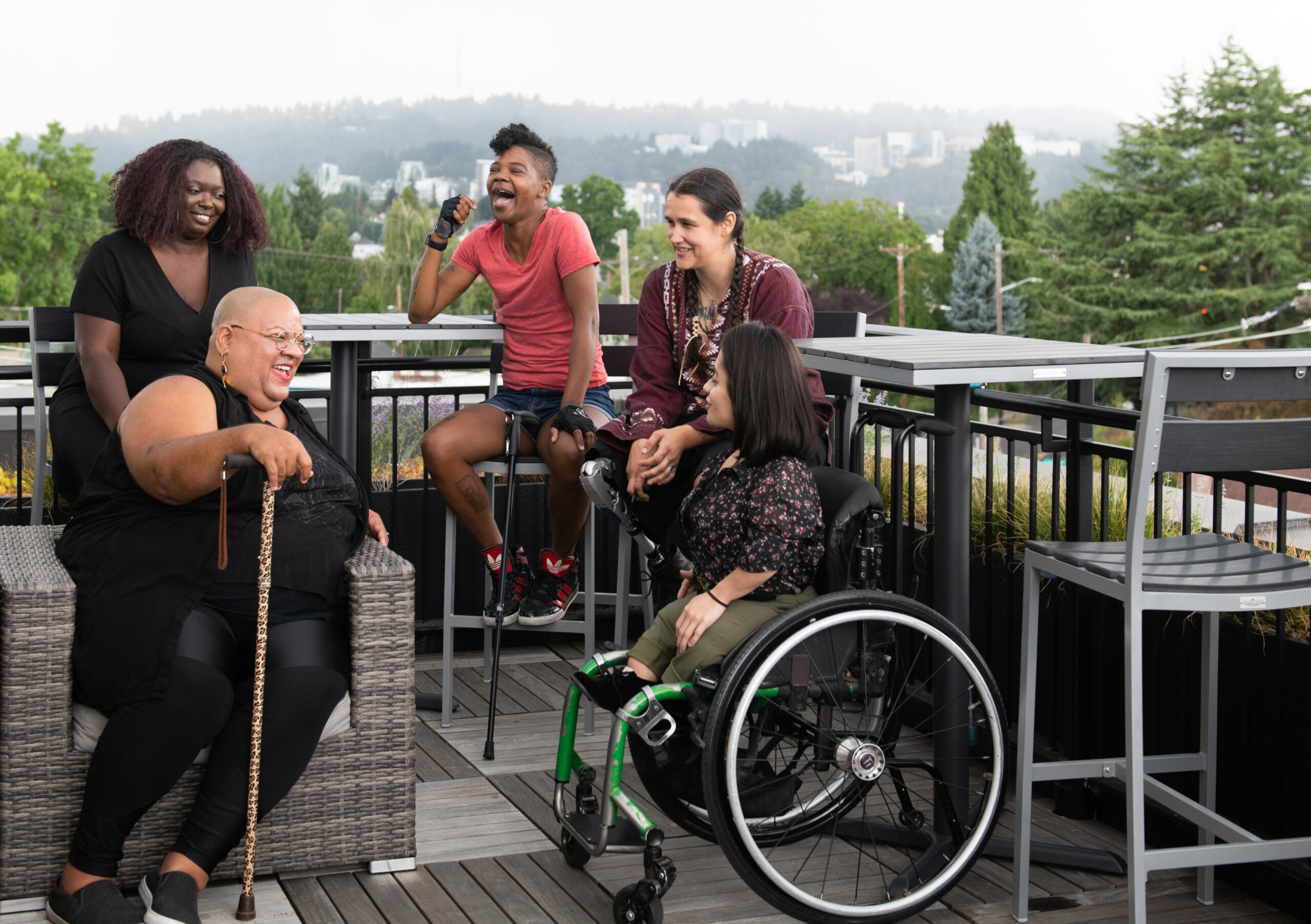 Five disabled people of color with canes, prosthetic legs, and a wheelchair sit on a rooftop deck, laughing and sharing stories.