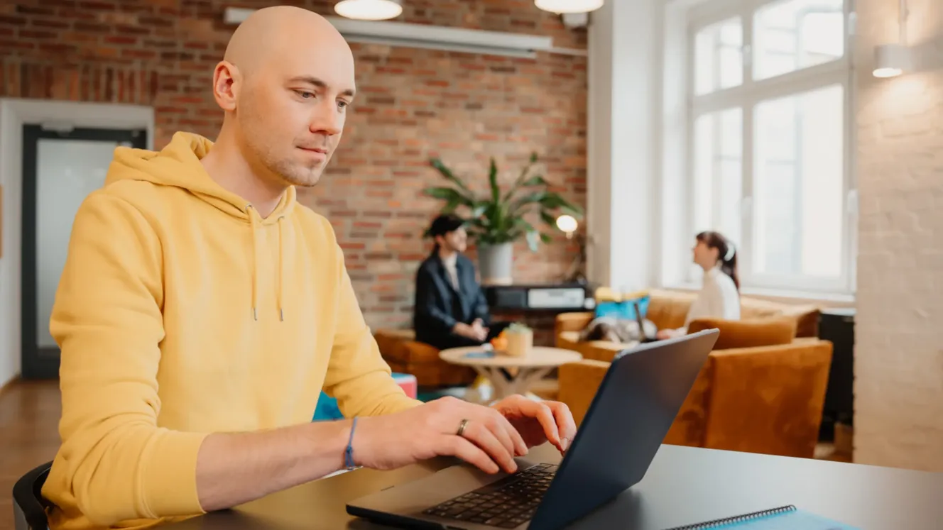 Staffbase employee typing on a laptop in a lounge area with a brick wall and colleagues chatting in the background.