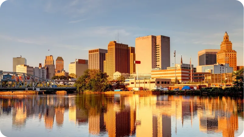 Newark, New Jersey skyline at sunset, reflected in the river with a bridge in the foreground.