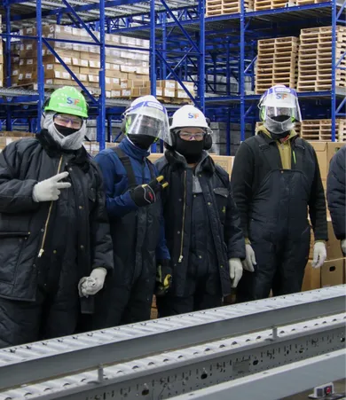 Four workers in protective gear and helmets stand in a warehouse with shelves of boxes in the background. Two of them are making peace signs.