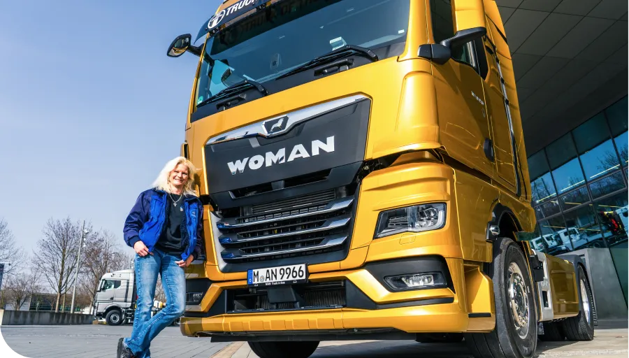 A person in a blue jacket leans against a large gold truck labeled "WOMAN" on the front, parked near a modern building.