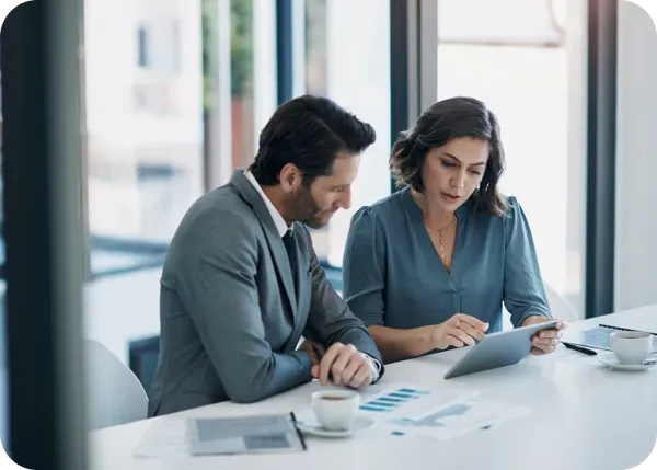 Two colleagues reviewing a tablet and charts at a bright office table with coffee cups.