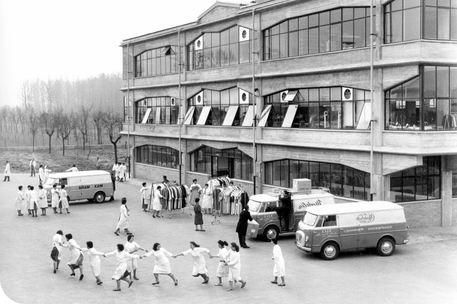 Black and white photo of a factory with workers in uniforms outside, some playing in a circle, and delivery vans parked nearby.