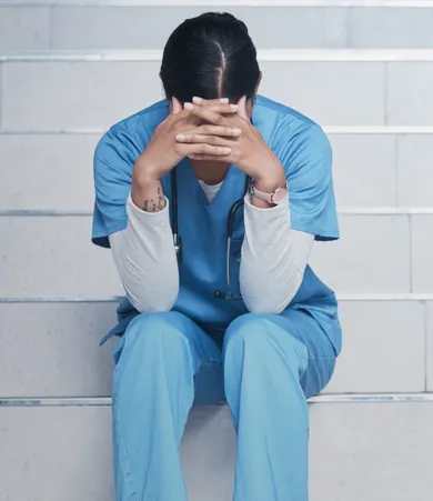 A person in blue scrubs sits on stairs, head in hands, appearing stressed. A stethoscope hangs around their neck.