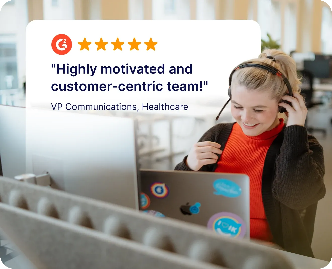 A smiling woman wearing a headset and red turtleneck works at a laptop covered in stickers in a modern open-plan office. Overlaid in the upper left is a G2 five-star review card with the quote: "Highly motivated and customer-centric team!" attributed to a VP of Communications in Healthcare.