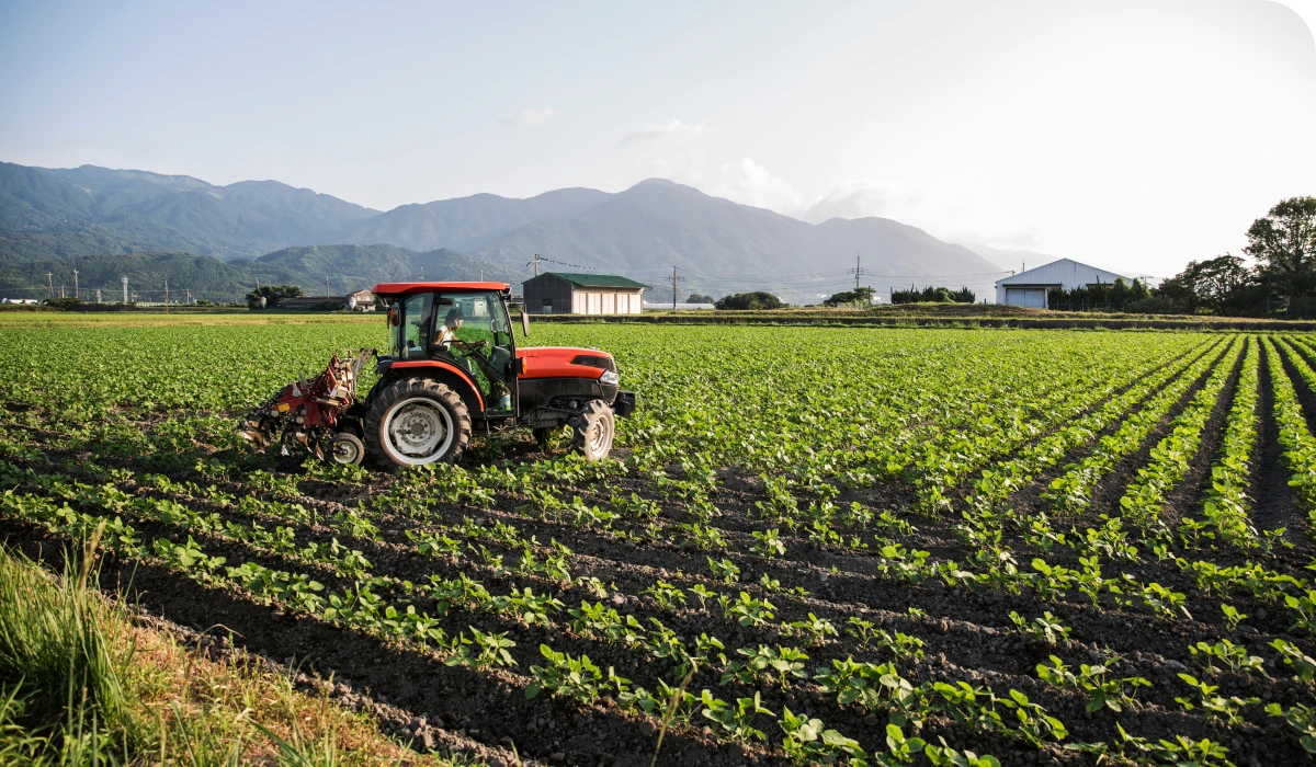 A red tractor plows a vast, green field with mountains in the background under a clear sky.