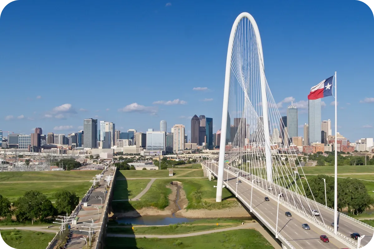 Aerial view of a modern white arch bridge with cars, a Texas flag, and a city skyline under a clear blue sky.