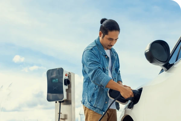 A man in a denim jacket charges an electric car at a charging station under a clear blue sky.