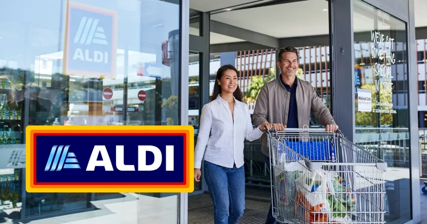 A smiling couple exits an ALDI store with a full shopping cart. The ALDI logo is prominently displayed.
