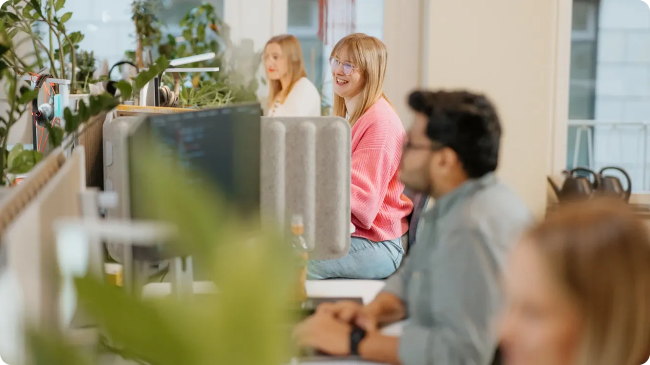 Staffbase team members chatting and smiling at their desks surrounded by office plants.