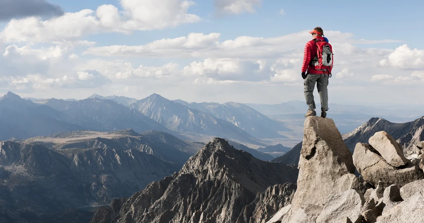 A person in hiking gear stands on top of a mountain and looks out over the summit.