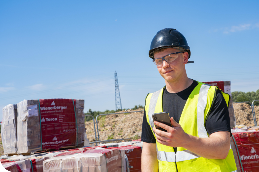 Construction worker in a hard hat and safety vest using a smartphone at a building site with brick stacks and blue sky in the background.
