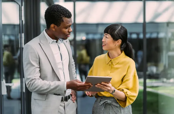 A man in a light gray suit and a woman in a yellow blouse are discussing something while looking at a tablet, standing in a modern office.