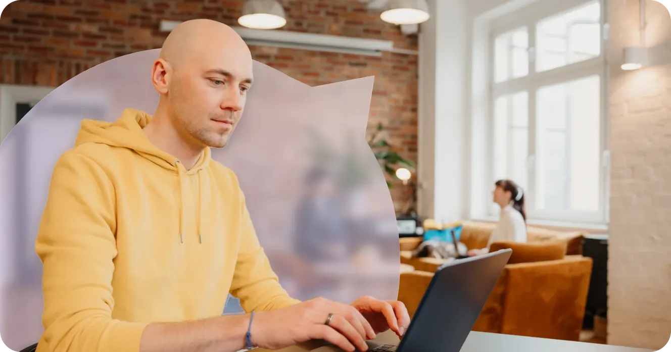Staffbase engineer typing on his laptop in a lounge area with a brick wall and colleagues in the background.