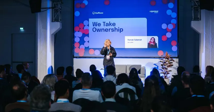 woman speaking on stage, Ramak Salamat, in front of audience, banner reading We Take Ownership