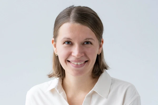 Smiling person with short brown hair, wearing a white shirt and small earrings, against a plain background.