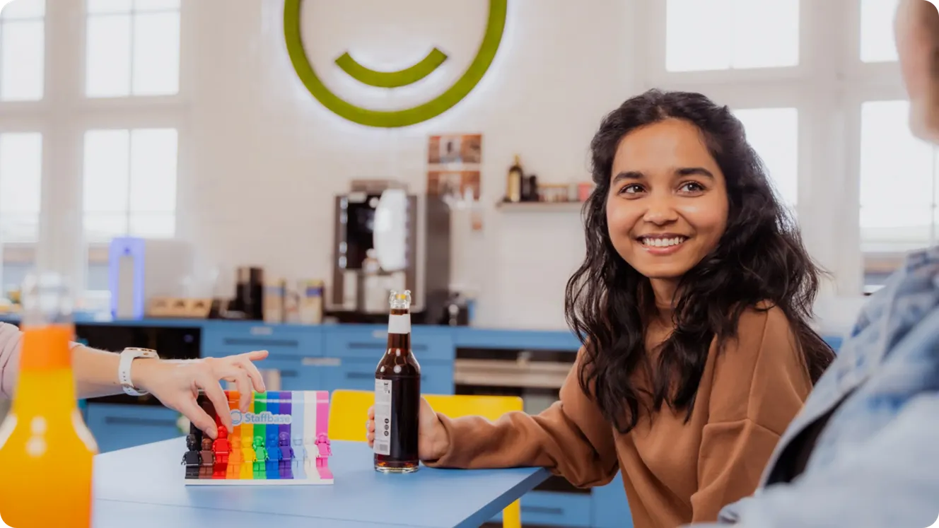 Staffbase employee smiling at a kitchen table with rainbow Lego figures and a Staffbase logo beneath the moss wall.
