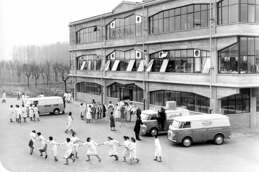 Black and white photo of a factory with workers in uniforms outside, some playing in a circle, and delivery vans parked nearby.