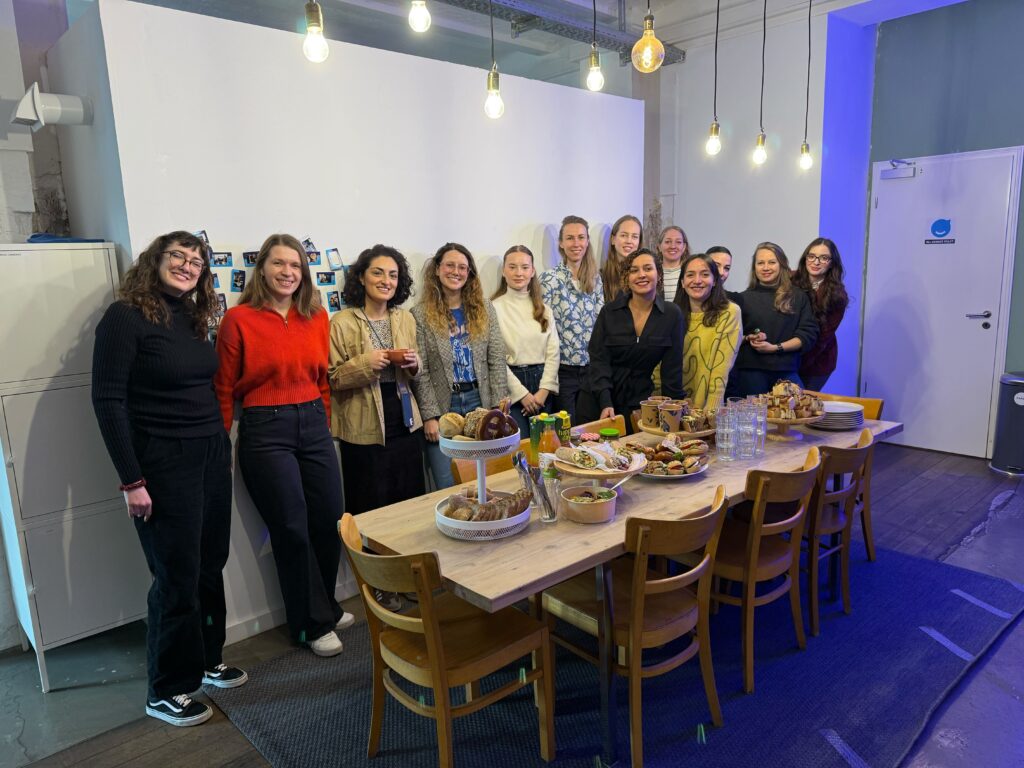 Group of about 13 women standing and smiling beside a long wooden table set with food and drinks in a room with string lights.