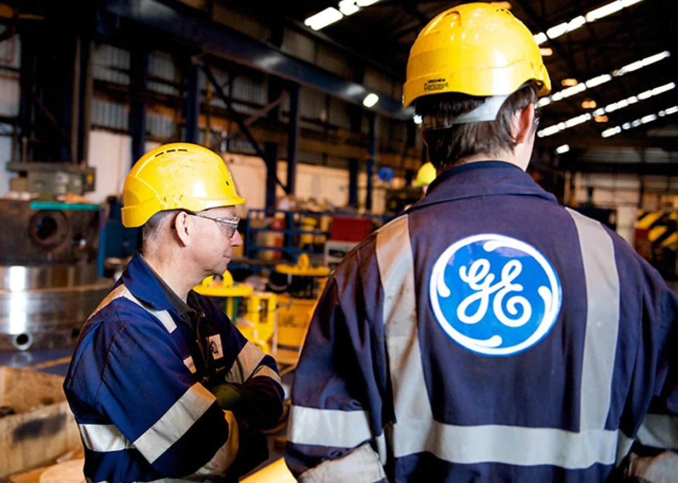 Two workers in blue uniforms and yellow helmets stand in an industrial facility. One has the GE logo on his back. Machinery is visible in the background.