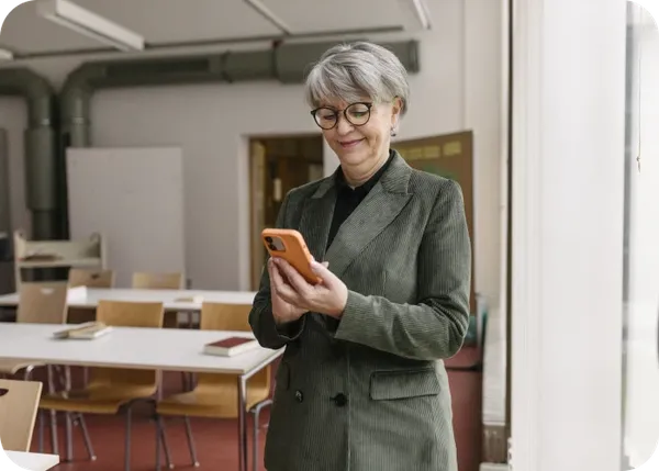 Smiling older woman in green blazer and glasses standing in a classroom, looking at an orange smartphone.