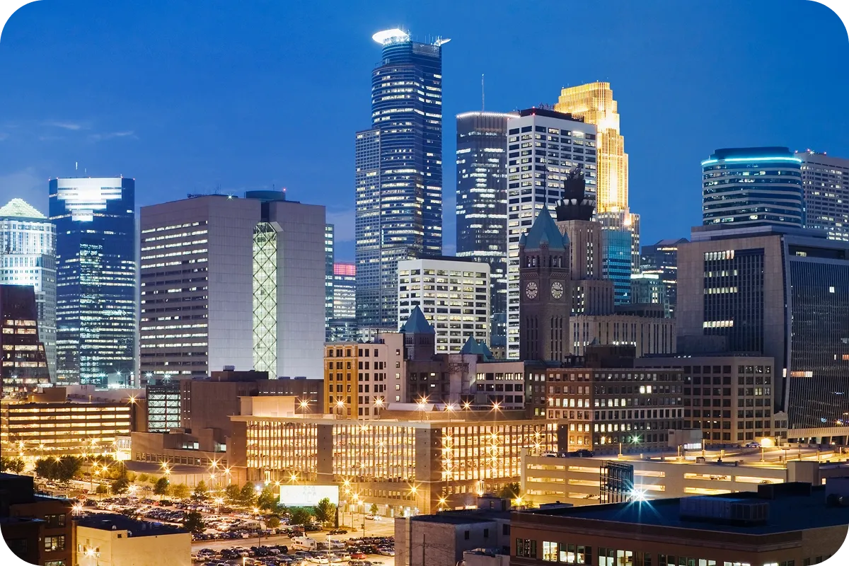 A vibrant city skyline at dusk with illuminated skyscrapers and a historic clock tower, set against a deep blue sky.