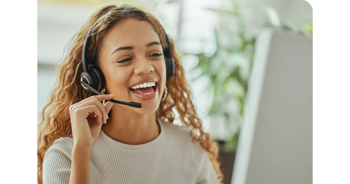Smiling woman with curly hair wearing a headset, sitting at a desk, engaging in a video call, with greenery in the background.