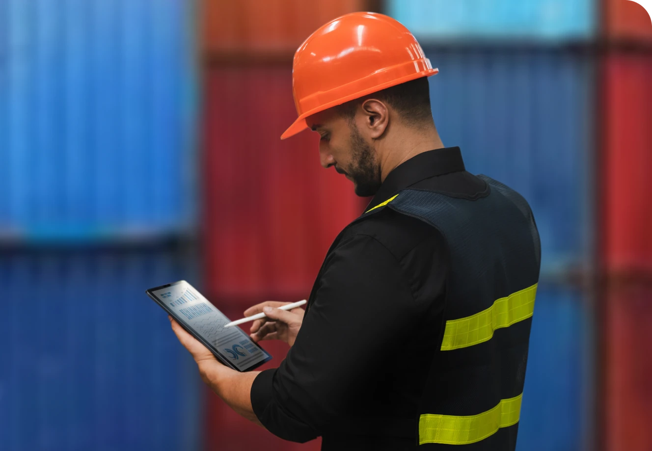 A man in a hard hat and reflective vest uses a tablet in front of stacked shipping containers.