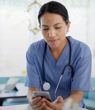 A healthcare professional in blue scrubs and a stethoscope, looking at a smartphone in a bright, clinical setting.