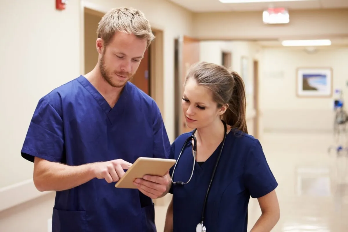 Two healthcare professionals in blue scrubs discuss information on a tablet in a hospital corridor.