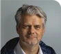 A middle-aged man with gray hair and a beard, wearing a white shirt and dark jacket, against a plain background.