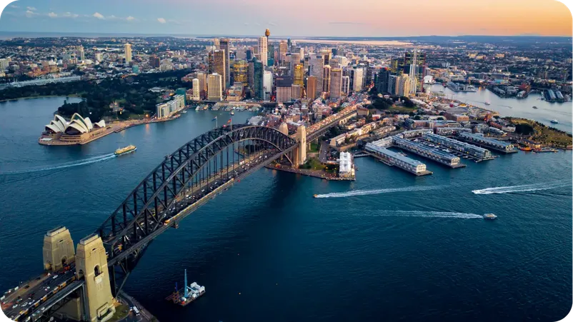 Aerial view of Sydney Harbour Bridge and the Sydney Opera House at sunset, with boats on the water.