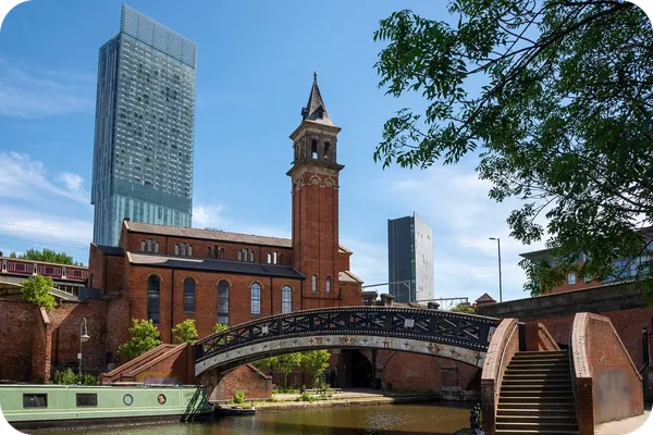 Historic brick church with a tall tower and arched bridge over a canal, set against modern skyscrapers under a clear blue sky.