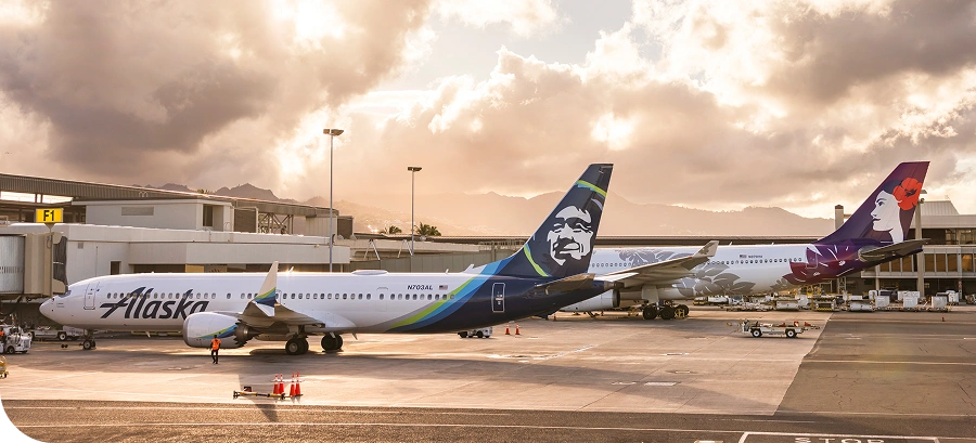 Two airplanes parked at an airport gate during sunset, with Alaska Airlines and another airline featuring a floral tail design.