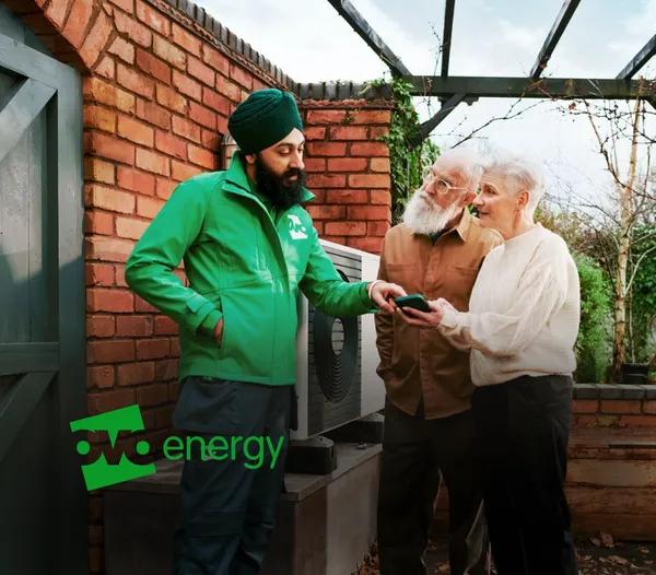 OVO Energy technician in a green jacket shows a smartphone to an older couple outside a brick home near an outdoor heating unit, with the OVO Energy logo overlaid