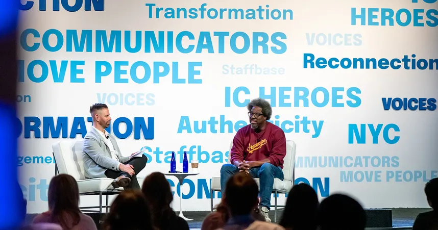Comedian, author, and documentary producer W. Kamau Bell sits on stage with VOICES conference host Adam Brayford. They are on stage with words like "transformation" and "reconnection" and phrases like "communicators move people" in blue text behind them on a white background. We see the backs of the audiences' heads listening to Kamau and Adam.