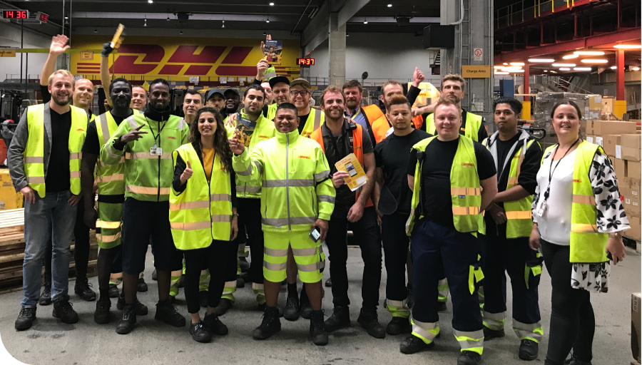 A diverse group of DHL employees in high-visibility clothing pose together in a warehouse, smiling and holding packages.