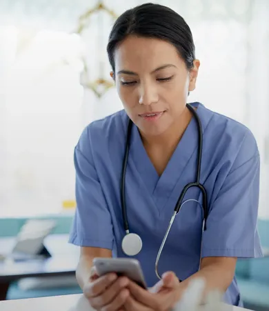A healthcare professional in blue scrubs and a stethoscope, looking at a smartphone in a bright, clinical setting.