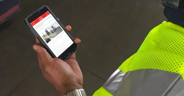 Person in a high-visibility jacket holding a smartphone displaying an app with a news article and image.