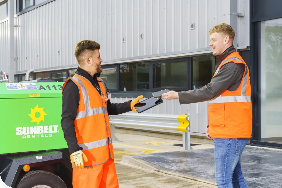 Two workers in high-visibility vests exchange a clipboard outside a Sunbelt Rentals building next to a green equipment unit.