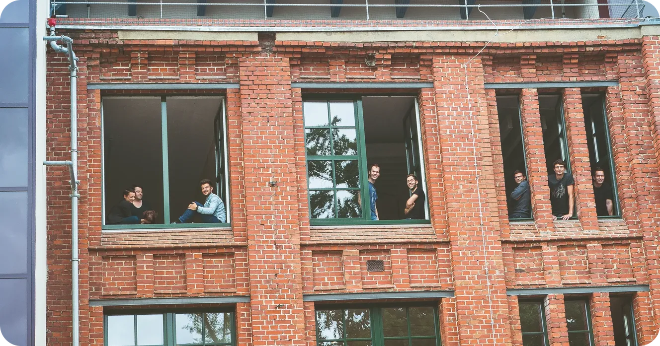 Staffbase employees smiling out of open windows in a red brick office building, including one with a dog.