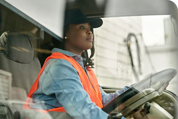 Woman in a safety vest and cap driving a vehicle, viewed through the windshield, focused on the road ahead.