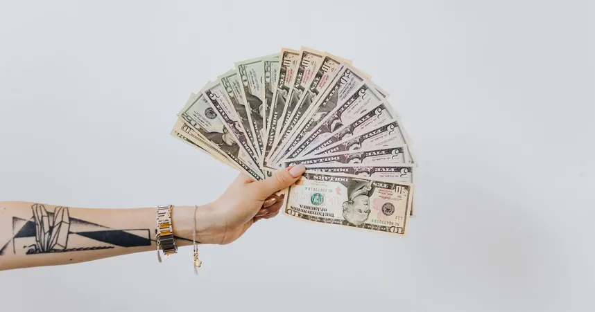 A hand fans out several American bills against a white backdrop. The person holding the money has bracelets and a long, geometric tattoo in black and white on their forearm.