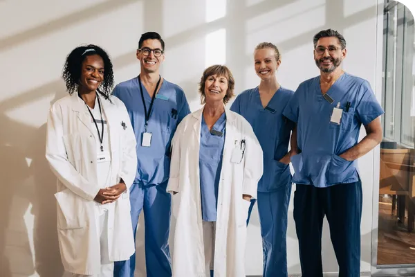 Five healthcare professionals in scrubs and lab coats smile while standing in a well-lit room, suggesting a medical or hospital setting.