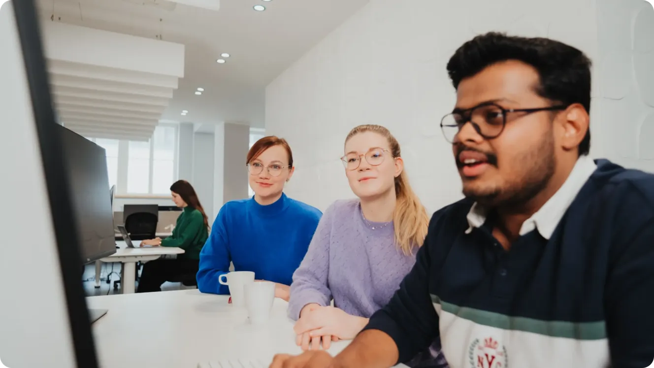 Three Staffbase colleagues smiling and talking together at a desk in a bright office. Title: Staffbase Colleagues Talking at Desk