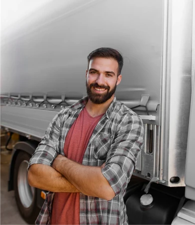 Smiling man with a beard, wearing a plaid shirt, stands confidently with arms crossed in front of a large truck.