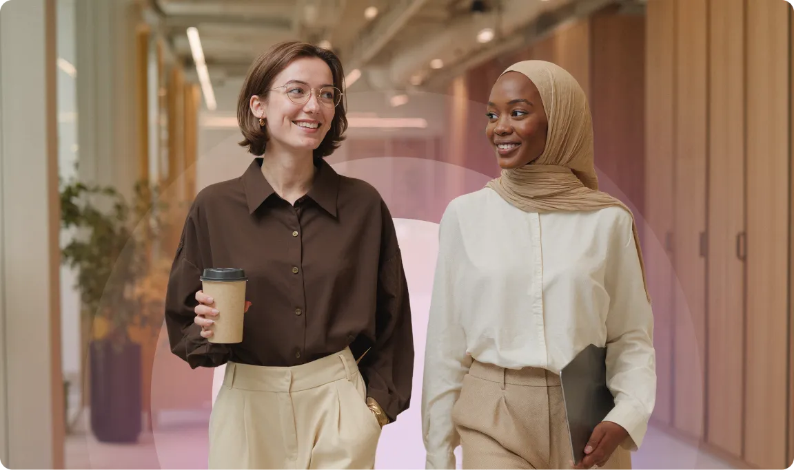 Two women walking in an office hallway, one holding a coffee cup. Both are smiling and dressed in professional attire.